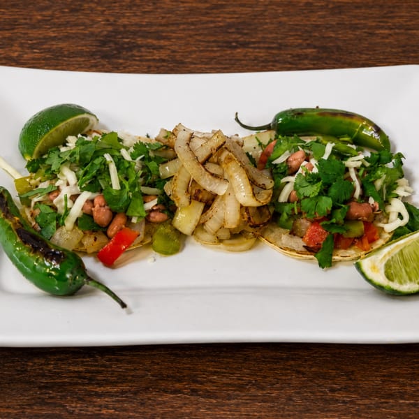 Three tacos with beans, cheese, cilantro, and grilled onions, garnished with limes and jalapeños.