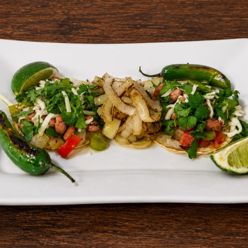 Three tacos with beans, cheese, cilantro, and grilled onions, garnished with limes and jalapeños.
