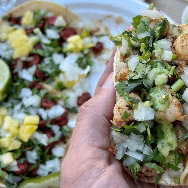 A hand holding a shrimp taco with lettuce and onions, with another taco in the background.