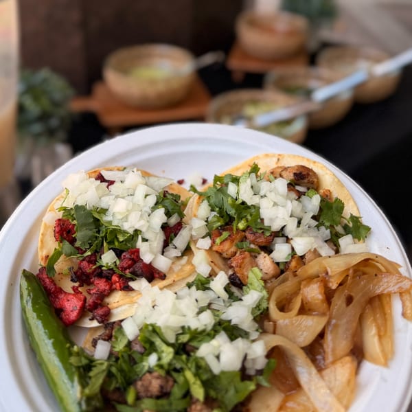 A white plate holds four street tacos with various fillings, onions, cilantro, and a green chili.