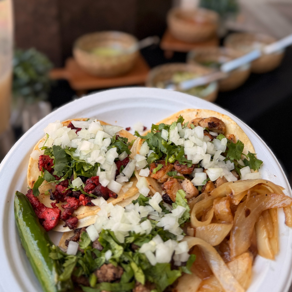A white plate holds four street tacos with various fillings, onions, cilantro, and a green chili.